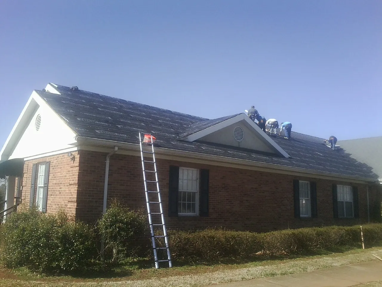 Crew tearing shingles off a brick ranch roof with a ladder set — Emergency Roof Repair work by our crew in Wingate